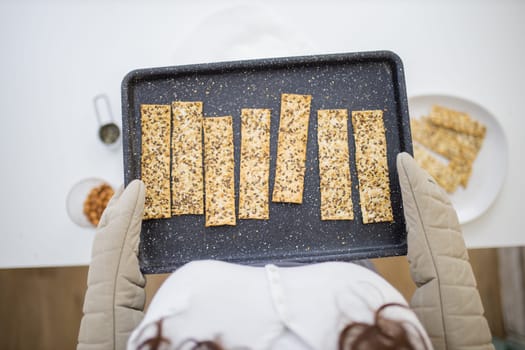 Woman with gloved hands holding black tray of Swedish multi-seed crispbread. Fresh crispy bread strips with linseed, amaranth, and rye from above. Healthy baked snacks
