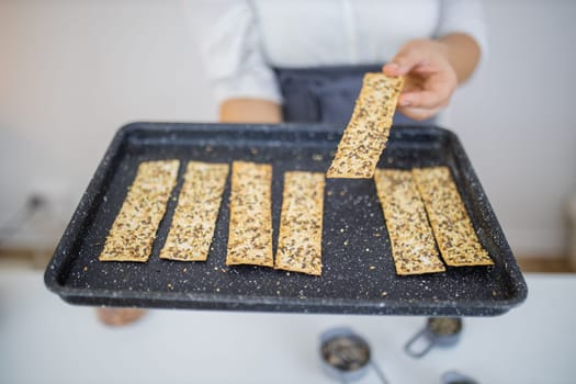 Female hand holding black tray of Swedish multi-seed crispbread. Freshly baked crispy bread strips with linseed, amaranth, and rye. Healthy baked snacks