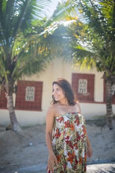 Beautiful woman wearing flowered dress standing in a tropical street with palm trees as background. Portrait of smiling brunette woman on sunny day. Tropical summer vacations