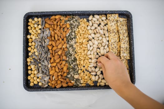 Female hand picking pistachios from black tray of chickpeas, almonds, peanuts, and multi-seed crispbread. Fresh crispy bread, seeds, and nuts from above. Healthy snacks