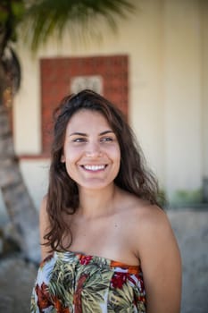 Beautiful woman wearing flowered dress standing in a tropical street with palm trees as background. Portrait of smiling brunette woman on sunny day. Tropical summer vacations