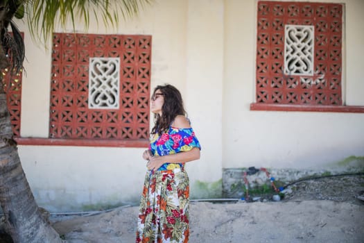Beautiful woman wearing flowered dress and sunglasses walking in a tropical street. Portrait of calm brunette woman on sunny day. Tropical summer vacations