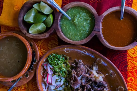 Top view of chopped lamb meat, hot sauces, lime slices, and lamb broth on colorful tablecloth. Tasty meal and condiments in clay plates and bowls above red and orange table. Classic Mexican cuisine