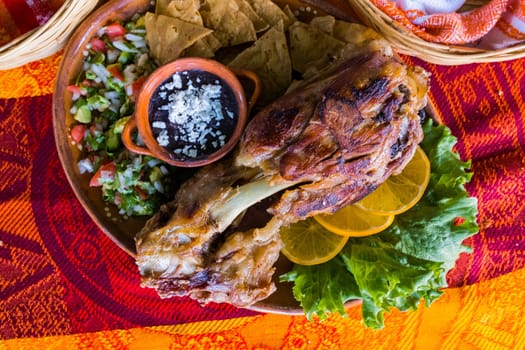 Top view of delicious roasted pork with lemon slices, lettuce, tortilla chips, and refried beans on colorful tablecloth. Tasty meat and accompaniments above orange and red table. Mexican style meal