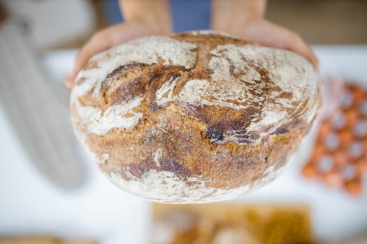 Female hands holding loaf of bread above blurry ingredients on table. Woman holding sourdough bread from above. Healthy meal preparation