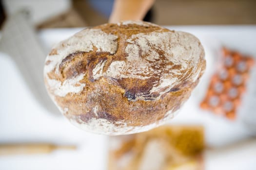 Female hand holding loaf of bread above blurry ingredients on table. Hand holding sourdough bread from above. Healthy meal preparation