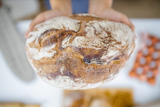 Female hands holding loaf of bread above blurry ingredients on table. Woman holding sourdough bread from above. Healthy meal preparation