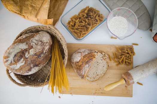 Bread, pasta, flour, and wooden cookware on white table from above. Ingredients and carbohydrates on cutting board and a basket. Healthy meal preparation