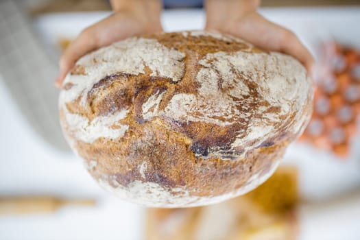 Female hands holding loaf of bread above blurry ingredients on table. Woman holding sourdough bread from above. Healthy meal preparation