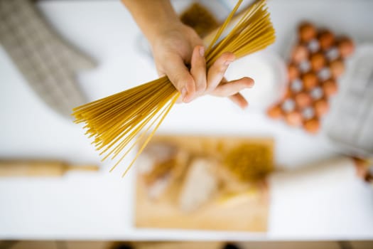 Female hand holding uncooked spaghetti above blurry ingredients on white table. Blurry eggs, pasta, flour, and bread on table from above. Pasta preparation