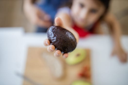 Happy little girl at white table showing hole avocado above wooden cutting board. Blurry young child making avocado toast. Kids preparing food