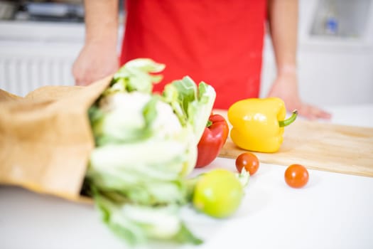 Paper bag of vegetables on white table and wooden cutting board. Man at table with cauliflower, bell peppers, lemons, and tomatoes. Healthy meal preparation
