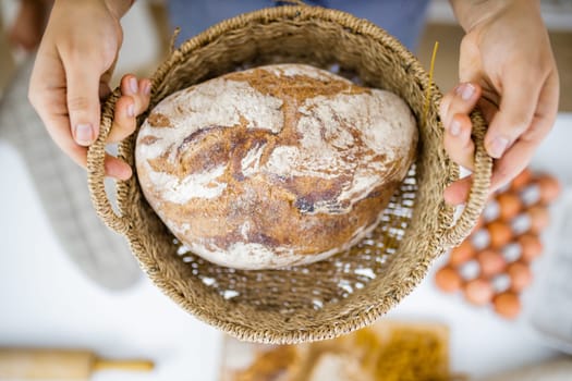 Female hands holding basket of sourdough bread above blurry ingredients. Blurry eggs, pasta, flour, and bread on table from above. Healthy meal preparation