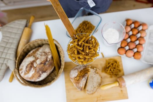 Uncooked fusilli on wooden pasta strainer above ingredients on white table. Eggs, spaghetti, flour, and bread on table from above. Pasta preparation