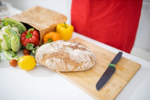 Bread on cutting board above white table and next to paper bag of vegetables. Sourdough bread alongside bell peppers, cauliflower, and lemons. Healthy meal preparation