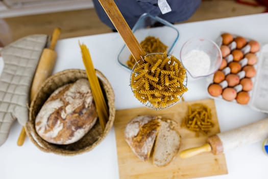 Uncooked fusilli on wooden pasta strainer above ingredients on white table. Eggs, spaghetti, flour, and bread on table from above. Pasta preparation