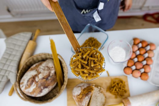 Uncooked fusilli on wooden pasta strainer above ingredients on white table. Eggs, spaghetti, flour, and bread on table from above. Pasta preparation