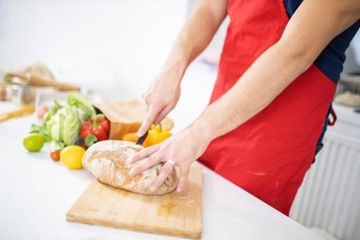 Male hands slicing bread on wooden cutting board above white table. Man cutting bread alongside bell peppers and cauliflowers. Healthy meal preparation