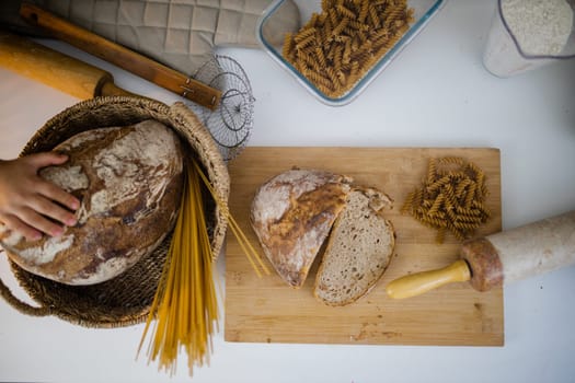 Bread, pasta, flour, and wooden cookware on white table from above. Ingredients and carbohydrates on cutting board and a basket. Healthy meal preparation
