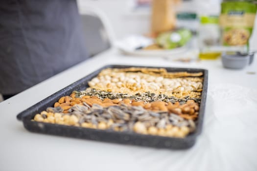 Black tray of chickpeas, almonds, peanuts, pistachios, and multi-seed crispbread. Fresh crispy bread, seeds, and nuts on white table. Healthy snacks