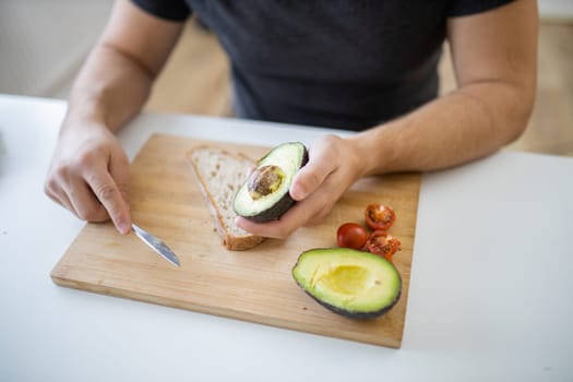 Male hand holding half an avocado above wooden cutting board with bread and tomatoes. Man at white table preparing avocado toast. Healthy meal preparation
