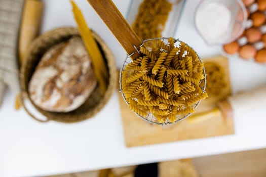 Uncooked fusilli on wooden pasta strainer above blurry ingredients on white table. Blurry eggs, spaghetti, flour, and bread on table from above. Pasta preparation