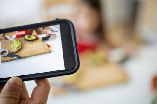 Hand with smartphone recording adorable little girl spreading avocado on slice of bread. Cute young child cutting vegetables on cutting board. Kids preparing food