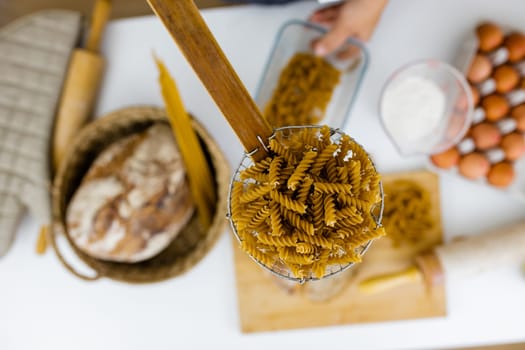 Uncooked fusilli on wooden pasta strainer above ingredients on white table. Eggs, spaghetti, flour, and bread on table from above. Pasta preparation