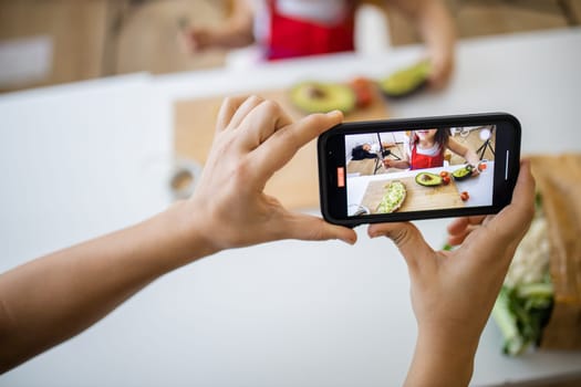 Female hands with smartphone recording adorable little girl spreading avocado on slice of bread. Cute young child cutting vegetables on cutting board. Kids preparing food