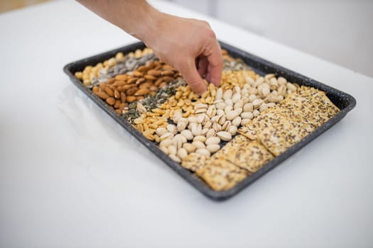 Male hand picking peanuts from black tray of chickpeas, almonds, pistachios, and multi-seed crispbread. Fresh crispy bread, seeds, and nuts above white table. Healthy snacks