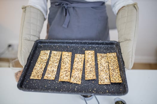 Gloved hands holding black tray of Swedish multi-seed crispbread. Freshly baked crispy bread strips with linseed, amaranth, and rye. Healthy baked snacks