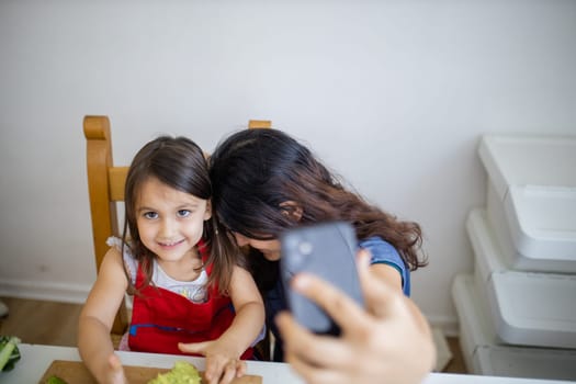 Happy mother and daughter taking selfie while eating toast with avocado. Cute young child cutting vegetables on cutting board. Mother-daughter cooking together