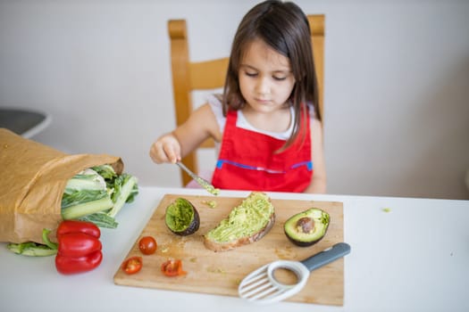 Adorable little girl at white table spreading avocado on slice of bread. Cute and concentrated young child cutting vegetables on cutting board. Kids preparing food