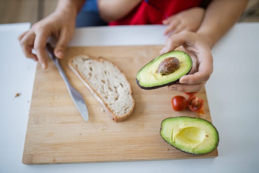 Male hands holding sliced avocado and knife above wooden cutting board. Man helping young child making avocado toast. Father-daughter cooking together