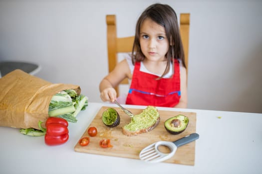 Adorable little girl at white table spreading avocado on slice of bread. Cute and concentrated young child cutting vegetables on cutting board. Kids preparing food