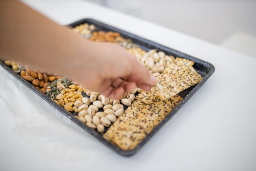 Female hand picking multi-seed crispbread from black tray of chickpeas, almonds, pistachios, and peanuts. Fresh crispy bread, seeds, and nuts above white table. Healthy snacks