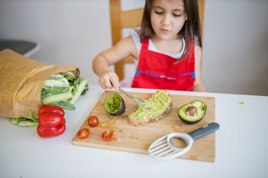 Adorable little girl at white table spreading avocado on slice of bread. Cute and happy young child cutting vegetables on cutting board. Kids preparing food