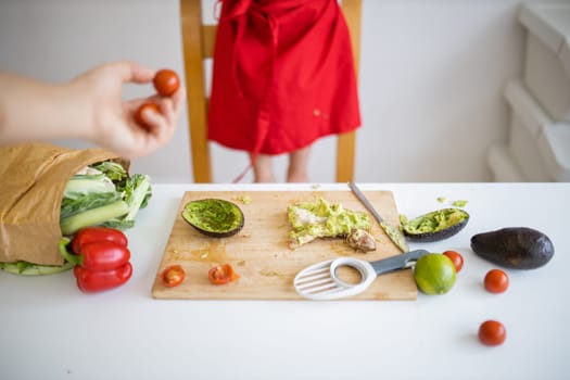 Female hand giving cherry tomatoes to little girl at a table making avocado toast. Young child cutting vegetables on cutting board. Kids preparing food