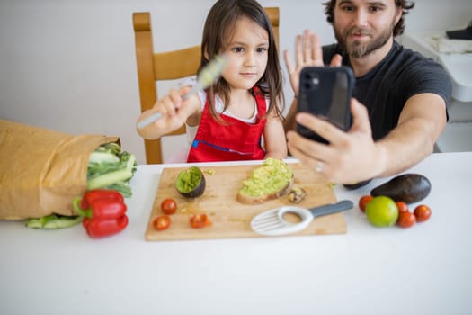Happy father and daughter taking selfie while spreading avocado on slice of bread. Cute young child cutting vegetables on cutting board. Father-daughter cooking together