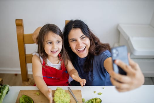 Happy excited mother and daughter taking selfie while eating toast with avocado. Cute young child cutting vegetables on cutting board. Mother-daughter cooking together