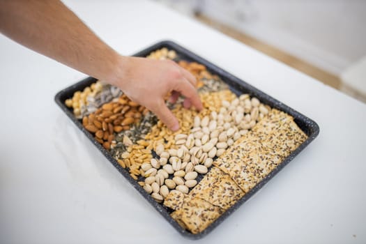 Male hand picking peanuts from black tray of chickpeas, almonds, pistachios, and multi-seed crispbread. Fresh crispy bread, seeds, and nuts above white table. Healthy snacks