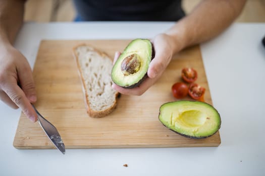 Male hand holding half an avocado above wooden cutting board with bread and tomatoes. Man at white table preparing avocado toast. Healthy meal preparation