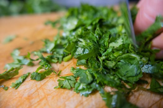 Close-up of hands chopping coriander on wooden cutting board. Person cutting green vegetables and herbs into pieces above wood surface. Traditional sauce preparation