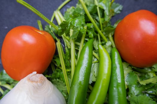 Close-up of three green chili peppers, onion, and tomato above coriander. Fresh green, red and white vegetables on dark blue surface. Healthy meal preparation
