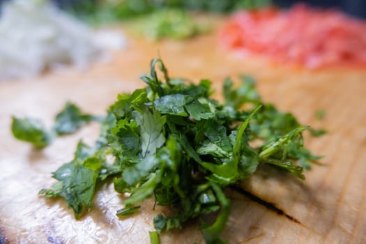 Close-up of chopped coriander on cutting board with tomato and chili pepper slices as background. Fresh green vegetables and herbs cut into pieces on wood surface. Traditional sauce preparation