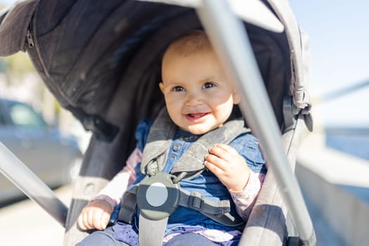 Adorable view of sunlight on happy baby sitting in gray stroller with blurry background. Portrait of cute smiling infant in blue dress during sunny day. Peaceful toddlers outdoors
