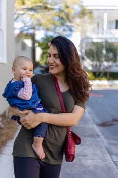 Portrait of happy mother holding her cute baby with blurry trees and houses as background. Smiling brunette woman and her adorable young daughter in street on sunny day. Lovely family outdoors