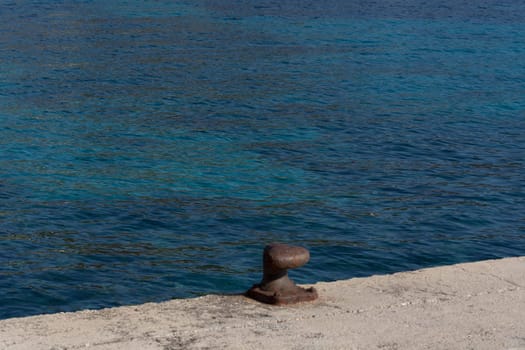 A rusty bollard on a quay wall with a rusty chain and in the background the water in the harbor on the Mediterranean Sea.