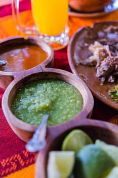 Close-up of brown bowls of lime slices and hot sauces, clay plate of chopped meat, and glass mug of juice. Tasty meal and Condiments above nice orange and red tablecloth. Classic Mexican cuisine