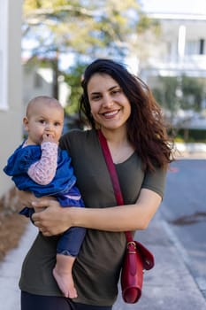 Portrait of happy mother holding her cute baby with blurry trees and houses as background. Smiling brunette woman and her adorable young daughter in street on sunny day. Lovely family outdoors
